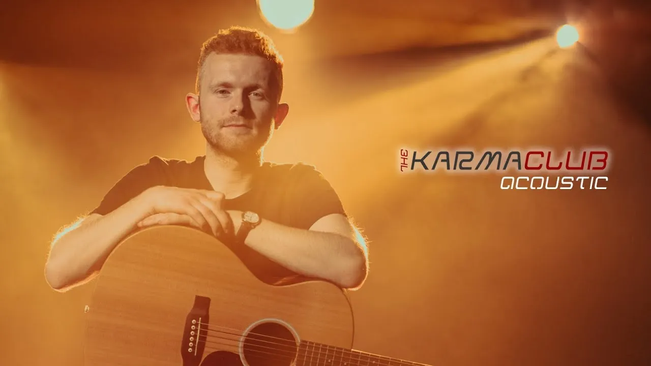 Man with short curly hair and a beard sitting with an acoustic guitar in warm stage lighting with 'The Karma Club Acoustic' text.