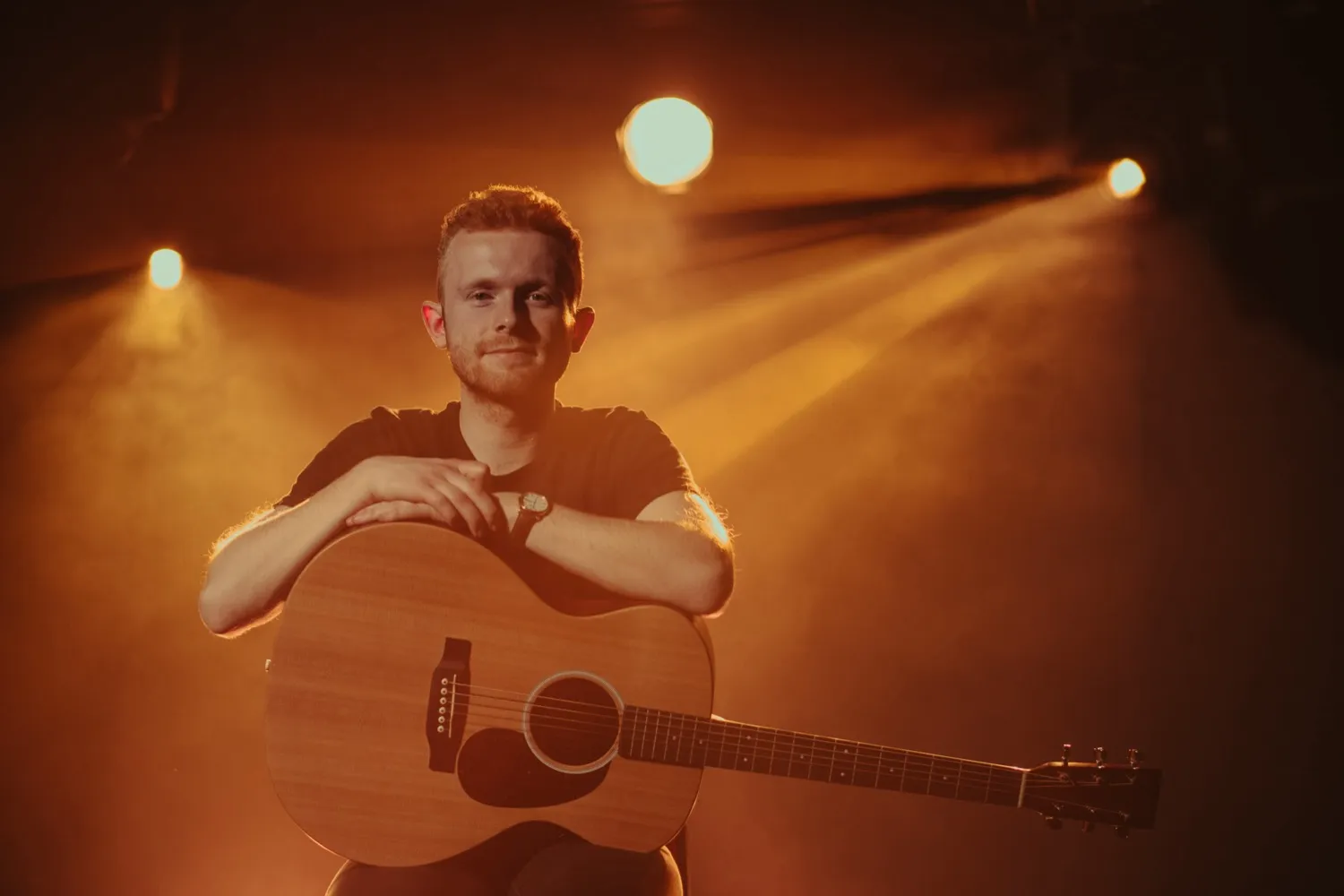 Man sitting with an acoustic guitar under warm stage lights and smoke.