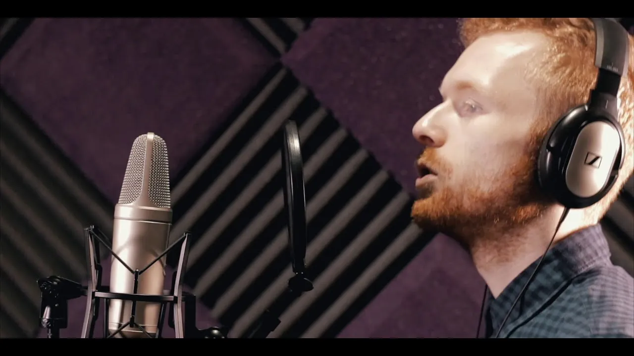 Red-haired man with headphones singing into a professional studio microphone with a pop filter in a soundproof room.