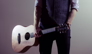 Person holding an acoustic guitar by its neck against a plain background.