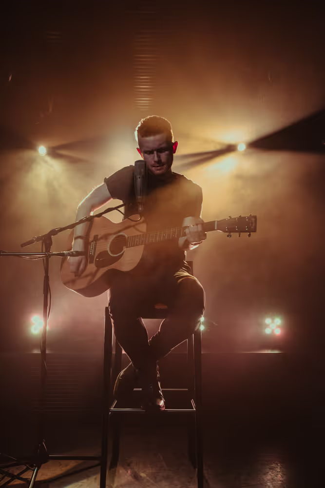 Man seated on a high stool playing an acoustic guitar with warm spotlights and haze in the background.