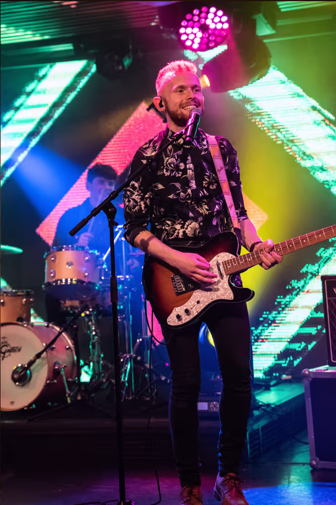Male musician smiling while playing electric guitar and singing on stage with drummer and colorful lights in background.